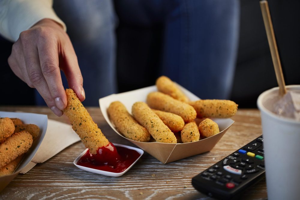 A hand dips a breaded mozzarella stick into marinara sauce on a wooden table, surrounded by more mozzarella sticks, a soft drink, and a TV remote.