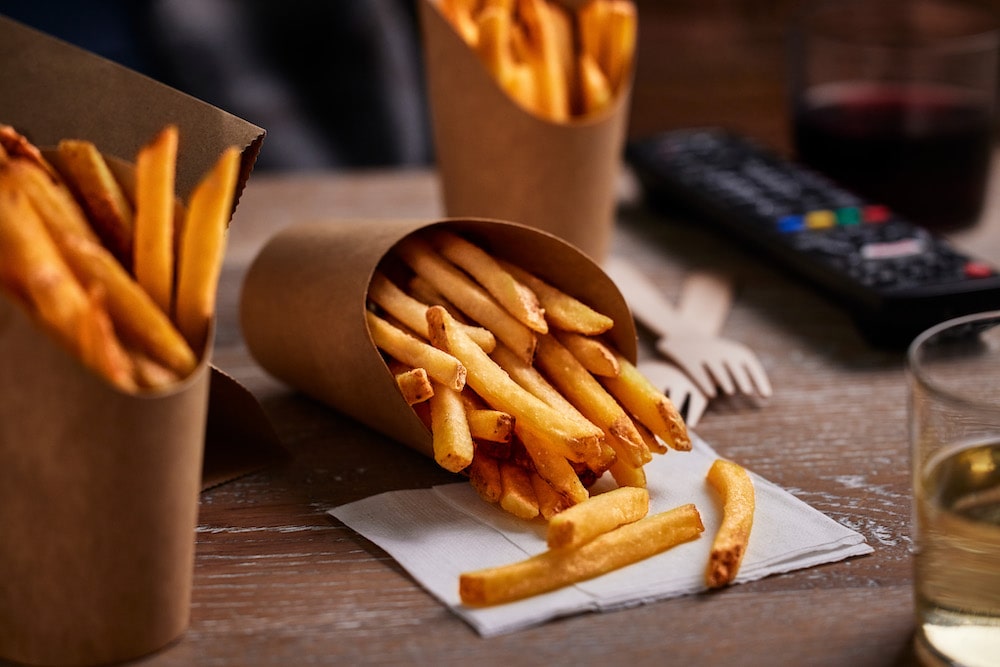 French fries spill from brown paper cones onto a wooden table, with drinks, a TV remote, and wooden forks creating a cozy snacking environment.