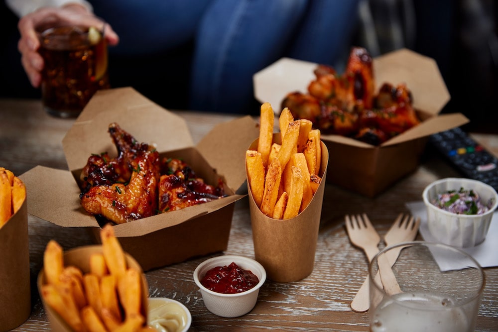Fried chicken wings and crispy fries sit in cardboard containers on a wooden table. Nearby, someone holds a drink, with sauces, utensils, and a remote in the background.