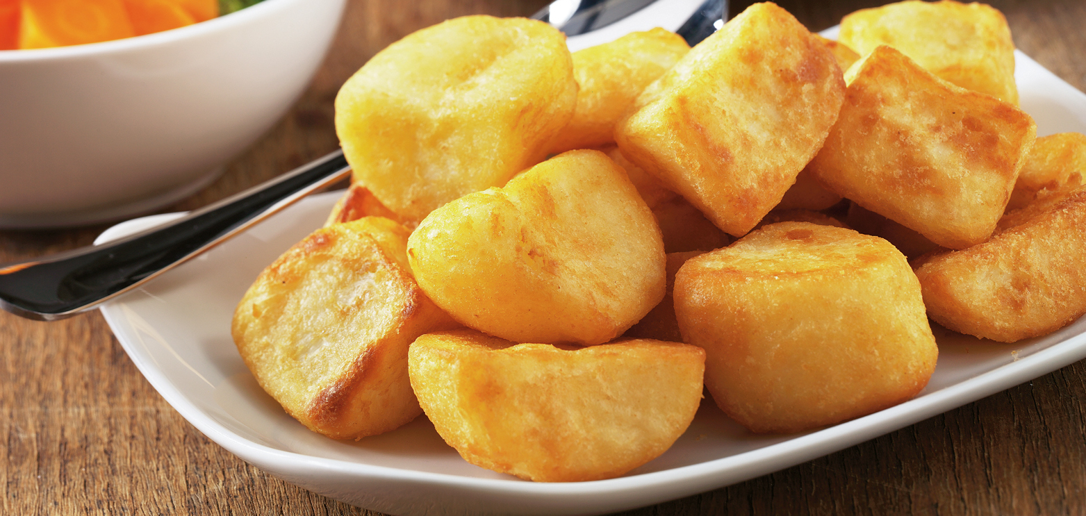 Golden, crispy roast potatoes rest on a white plate with a fork, set on a wooden table. A bowl of sliced vegetables is partially visible in the background.