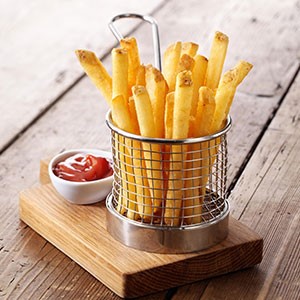 Golden french fries rest in a small metallic basket on a wooden board, accompanied by a small white dish of ketchup, on a rustic wooden table.