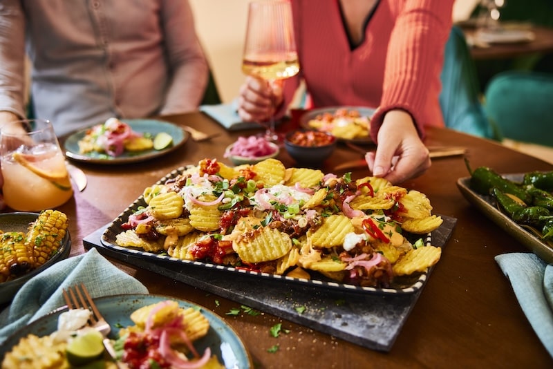 A platter of loaded McCain Deep Ridge Slices, topped with sour cream, onions, and garnish, is being reached for on a dining table surrounded by people with drinks and side dishes.